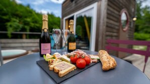Una mesa con diversos alimentos, como pan, tomates y una botella de vino, ubicada al aire libre con una estructura de madera al fondo.