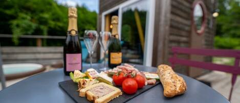 Un tavolo imbandito con diversi cibi, tra cui pane, pomodori e una bottiglia di vino, all'aperto con una struttura in legno sullo sfondo.