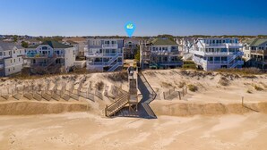 A coastal residential area with multiple two-story houses, a central staircase leading to a beach, and a clear blue sky. - The beautiful beaches of Corolla are just a few feet away at Oasis! (Corolla)