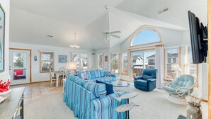 A spacious living room with a large sofa, a blue armchair, and a round coffee table. There is a dining area with a table and chairs, and a television mounted on the wall. - The beautiful beaches of Corolla are just a few feet away at Oasis! (Corolla)