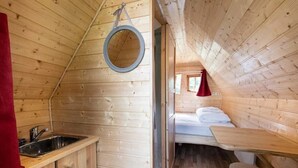 A wooden cabin with a kitchen area featuring a sink and cabinets, a small window, and a bed with a red curtain. - ZEcamping 2 étoiles piscine couverte animaux acceptés locatif 6m2 2 pers (Bar-sur-Aube)