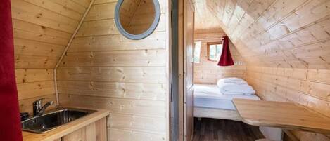 Una cabaña de madera con una zona de cocina que incluye un fregadero y armarios, un ventanal pequeño y una cama con cortina roja.