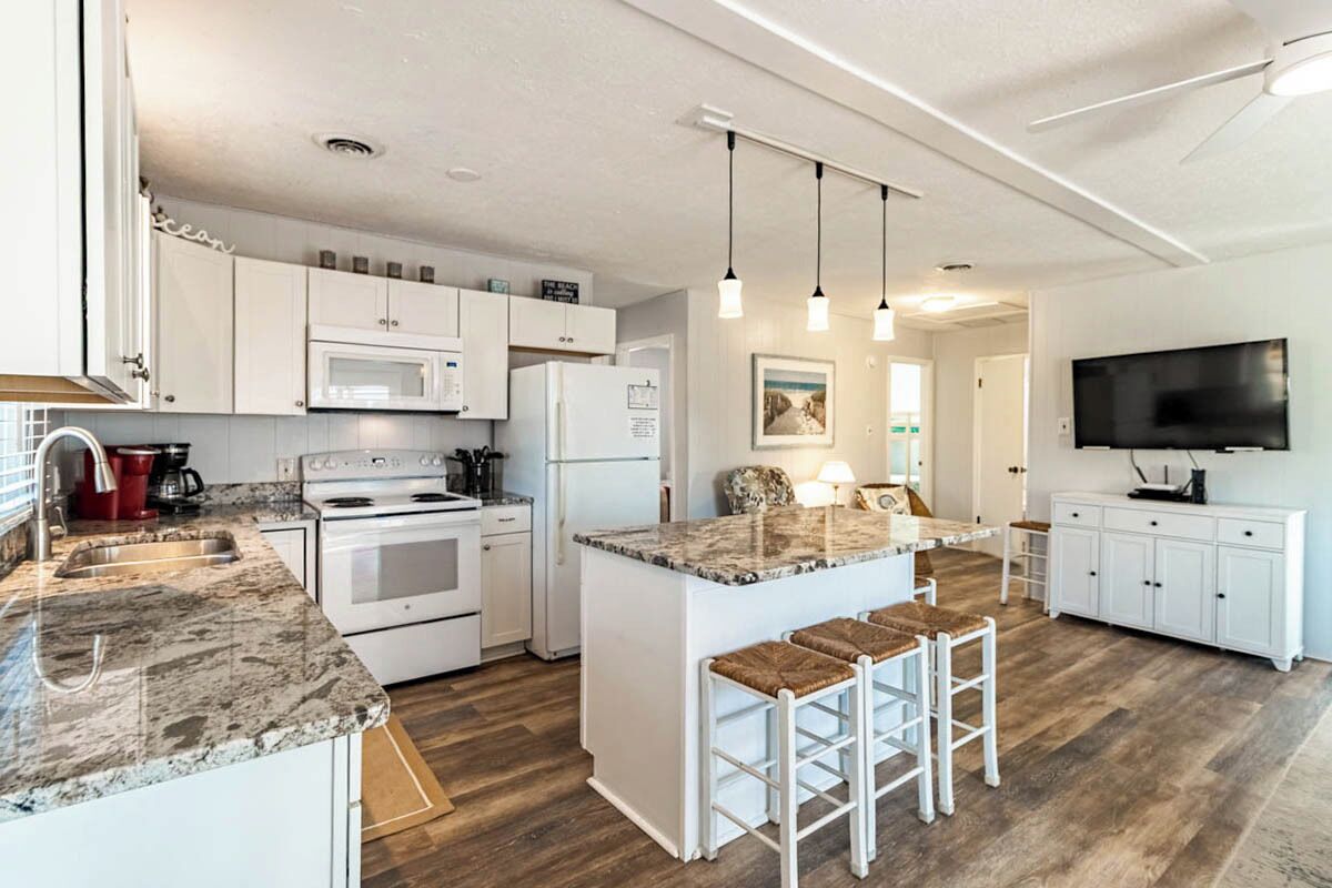 A modern kitchen with white cabinets, a granite countertop, a central island with bar stools, a microwave, a refrigerator, and a flat-screen TV.