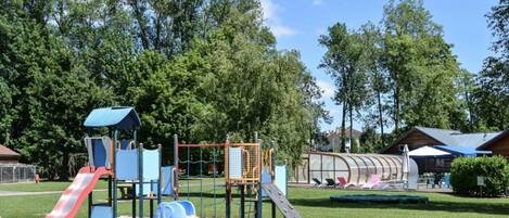 A playground with slides and climbing structures, surrounded by trees and a covered seating area.