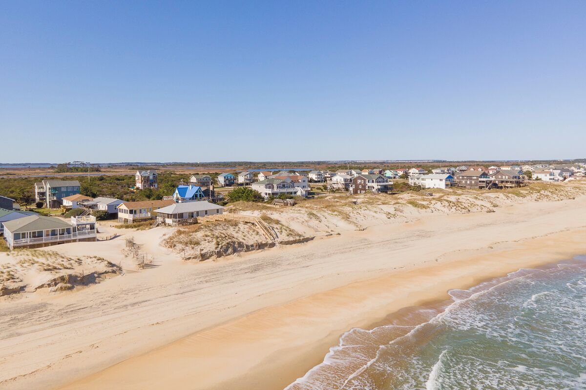 Una zona residencial costera con casas a lo largo de la playa, dunas de arena y cielo despejado.