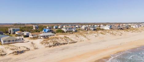 Una zona residencial costera con casas a lo largo de la playa, dunas de arena y cielo despejado.