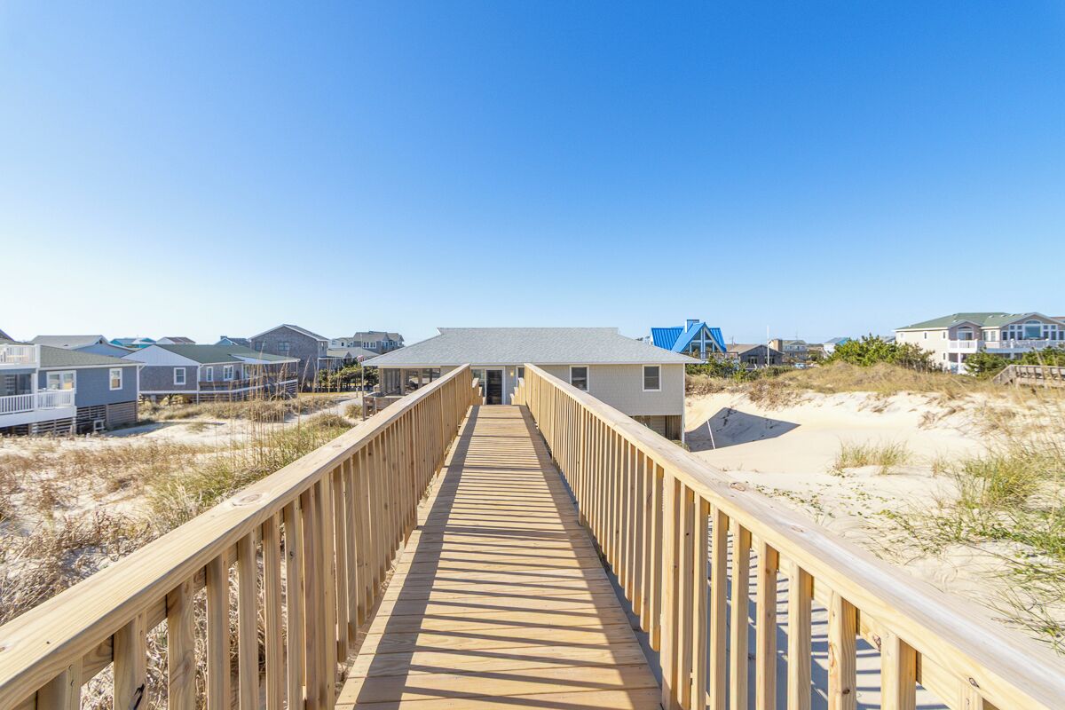 A wooden boardwalk leading to a beach house with a grey roof, surrounded by other beach houses.