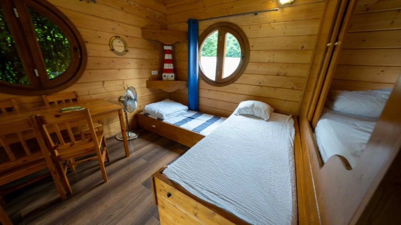 Eine Holzhütte mit rundem Fenster, einem kleinen Tisch und einem Bett mit karierter Bettdecke.