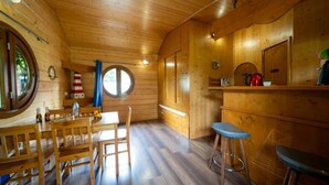 Interior de una cabaña de madera con comedor, barra y ventanal circular.