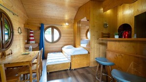 Interior de una cabaña de madera con comedor, dos camas individuales, una pequeña barra y un ventanal circular.