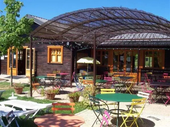 An outdoor seating area with tables and chairs, a wooden building with windows, and a covered patio.
