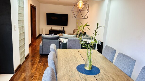 A modern living room with a wooden dining table, grey sofas, and a TV mounted on the wall.