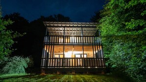 Une cabane en bois à deux étages, dotée de grandes fenêtres et entourée d’une végétation luxuriante.
