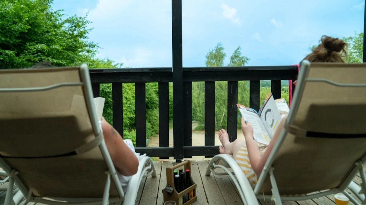 Twee loungestoelen gericht op een houten terras met een reling, een kleine tafel met flessen, en groen in de achtergrond.