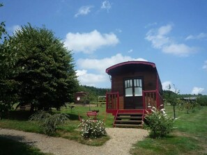 Una cabaña de madera roja con un porche y un camino de grava que la conduce, rodeada de vegetación y árboles.