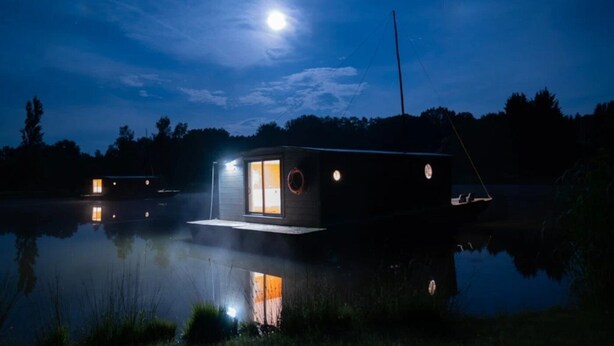 Een klein, modern vakantiehuisje aan een rustig meer 's nachts, met een zichtbare maan aan de hemel.