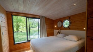 Een slaapkamer met een groot raam, houten wanden, een bed met witte lakens en twee spiegels aan de muur.