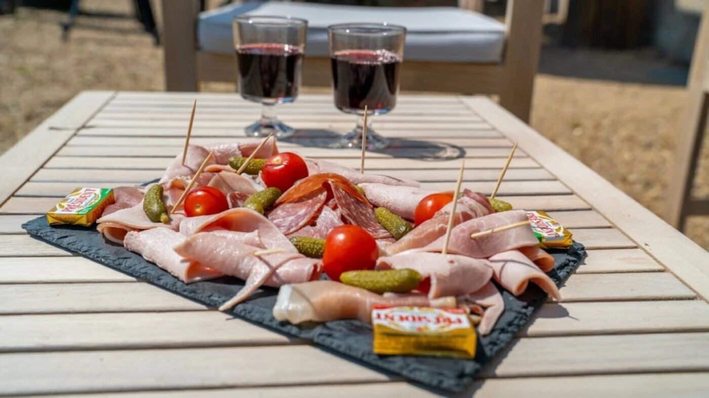 Une table avec une planche de charcuterie, deux verres de vin rouge et une chaise en arrière-plan.