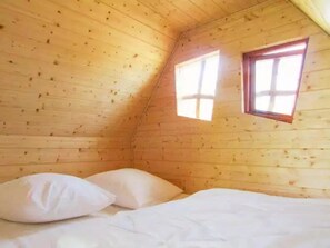 Une petite pièce chaleureuse aux murs en bois et au plafond en pente. On y trouve un lit simple avec une literie blanche et deux oreillers. Une petite fenêtre à cadre en bois laisse entrer la lumière naturelle dans la pièce.