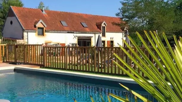 A swimming pool with a wooden fence, a house with a sloped roof, and a clear sky. - ZEcamping piscine chauffée animaux acceptés sauna cabane BBQ 6m2 (Chassenard)