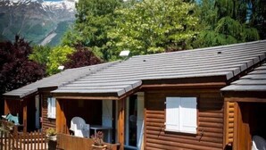 A row of wooden cabins with a gravel driveway, surrounded by greenery and a mountain backdrop. - ZEcamping 4 étoiles piscine couverte animations chalet TV 35m2 5 pers (Le Bourg-d'Oisans)