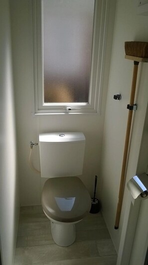 A small, compact bathroom with a toilet, a window, and a wooden shelf.