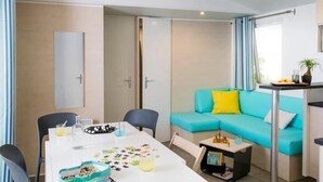 A modern living room with a teal sofa, a white dining table, and a small side table with a plant. - ZEcamping 3 étoiles parc aquatique chiens acceptés locatif TV clim 30m2 (Frejus)