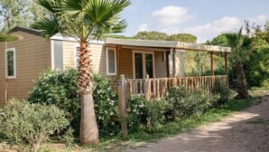 A single-story mobile home with a covered porch, surrounded by lush greenery and a dirt path. - ZEcamping 3 étoiles parc aquatique chiens acceptés locatif TV clim 30m2 (Frejus)