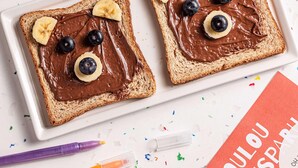 Twee sneetjes toast met chocoladesiroop, bananen en blauwe bessen, gerangschikt om ogen en een mond voor te stellen.
