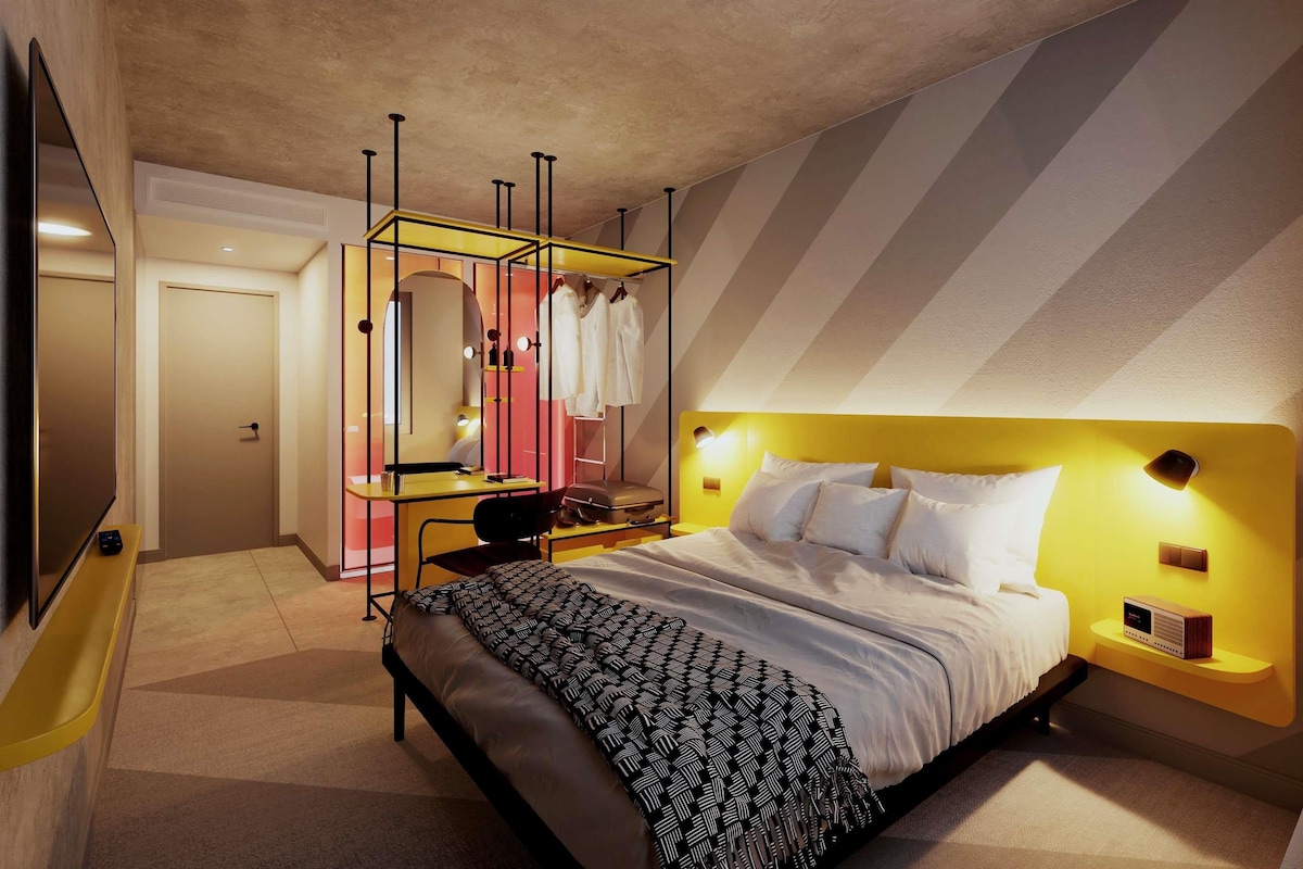 Une chambre d’hôtel moderne équipée d’un lit, d’une table de chevet et d’un coin coiffeuse avec un miroir et un porte-vêtements.