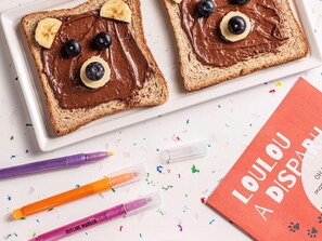Two slices of toast with chocolate spread, bananas, and blueberries arranged to resemble eyes and a mouth. - Ibis Styles Hamburg Hafencity (Hamburg)