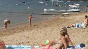 Een strand met mensen die zonnebaden, een zeilboot en diverse andere boten op het water.