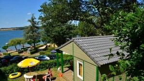 Une petite cabane en bois, avec un toit recouvert de tuiles, entourée d’une végétation luxuriante et bordée par un plan d’eau.