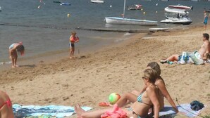 Ein Strand mit Menschen, die sonnenbaden und spielen, Booten im Wasser und einem klaren Himmel.
