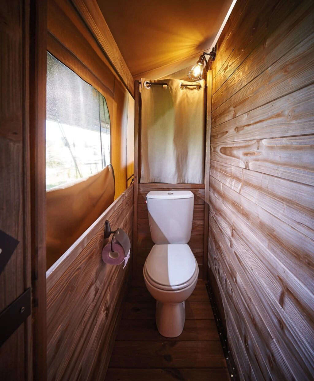 A narrow wooden cabin bathroom with a toilet, a window, and a towel rack.