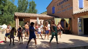 Un groupe de personnes danse à l’extérieur, devant un bâtiment sur lequel on peut lire l’inscription : « CAMPING La Rivière : BAR À SNACKS ET PIZZERIA ».