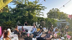 Een terras met ronde tafels en stoelen, omgeven door groen en bloemen.