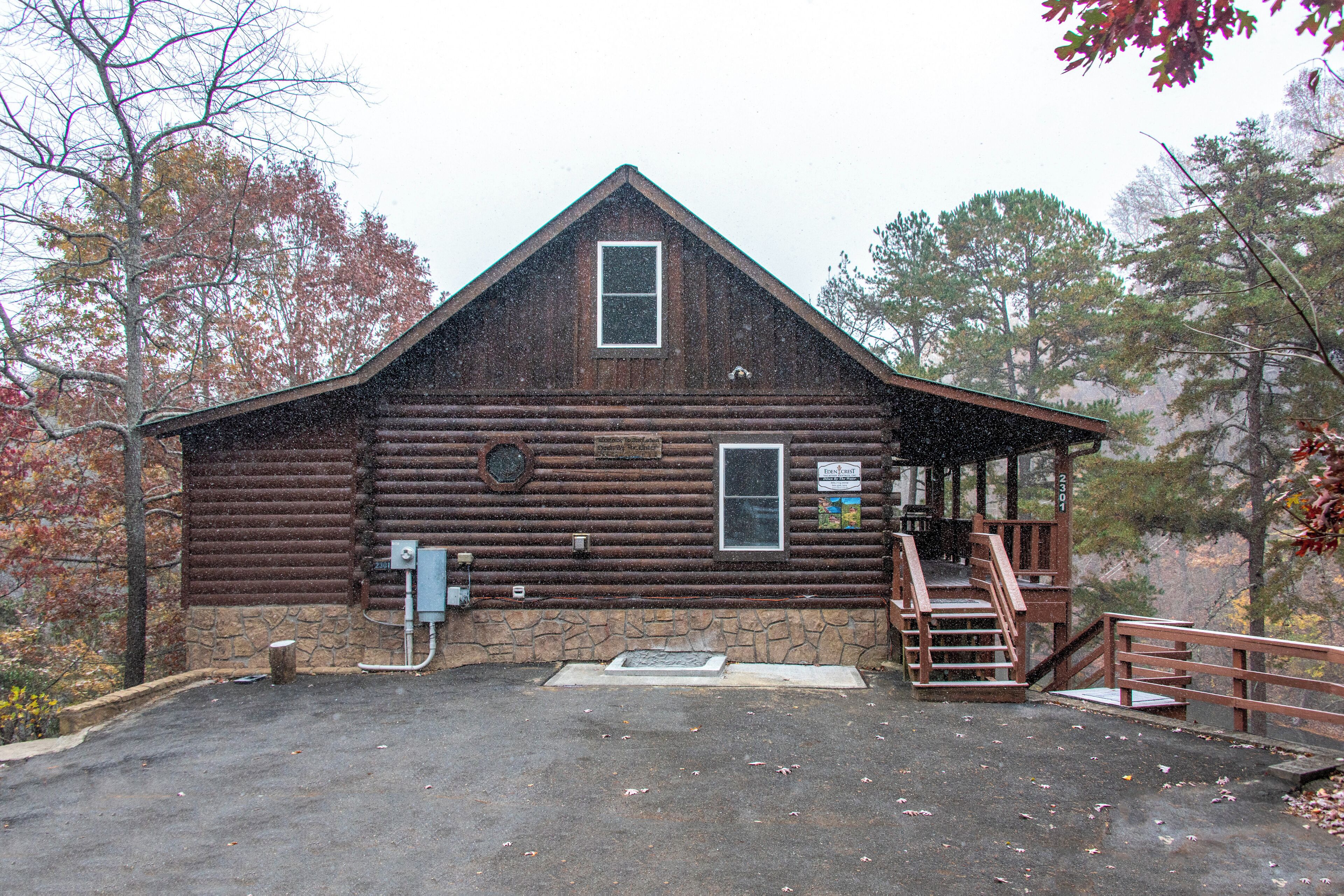 A rustic log cabin with a stone foundation, a front porch with a wooden railing, and a small staircase leading to a second-story deck.