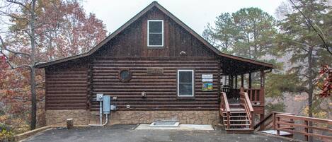 A rustic log cabin with a stone foundation, a front porch with a wooden railing, and a small staircase leading to a second-story deck.