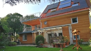 Una casa de madera de dos pisos con balcón, paneles solares y un perro en el jardín.