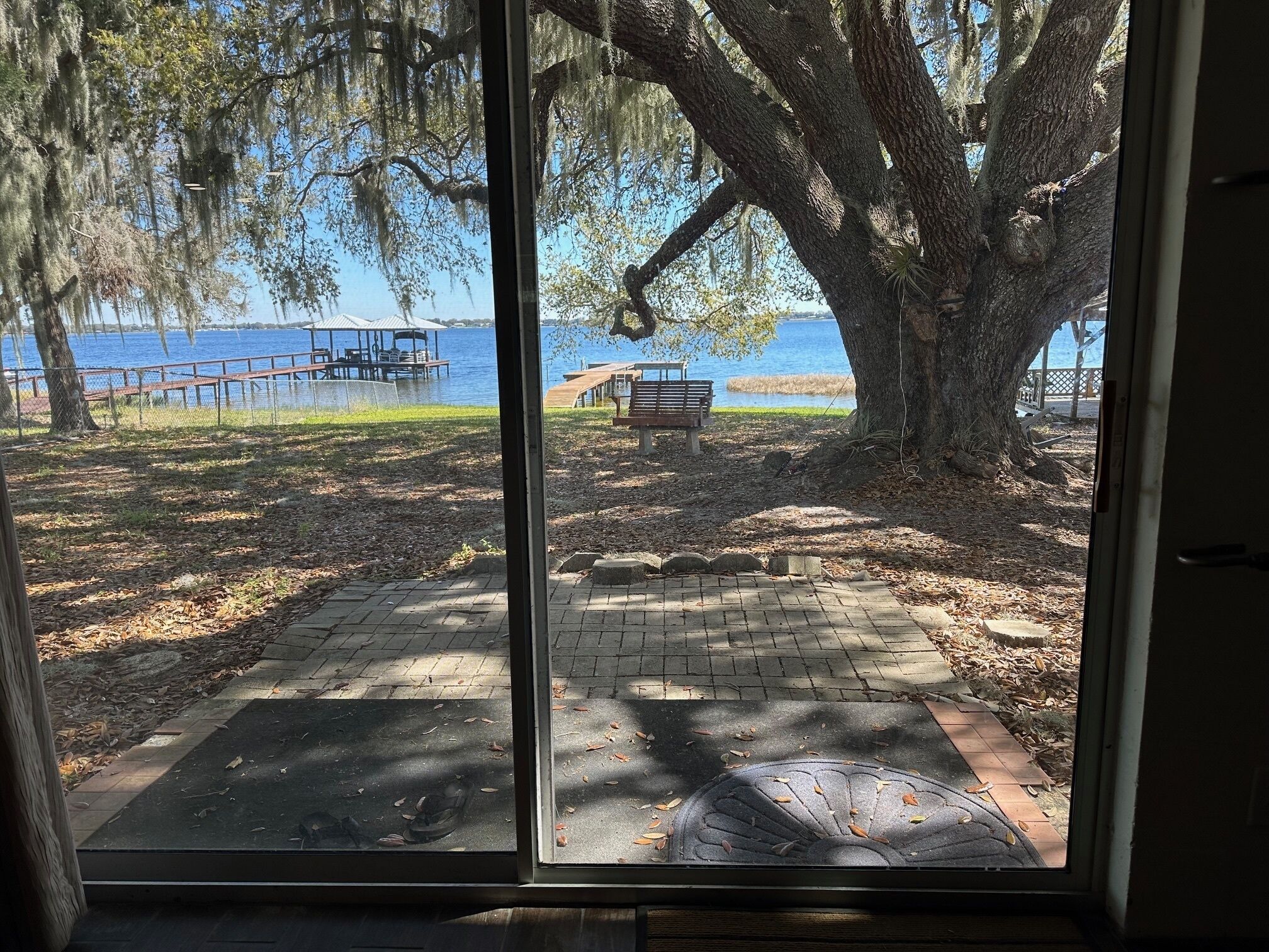 A view through a glass door of a park with a bench, a paved area, and a body of water in the background.