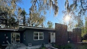 Una casa moderna de dos pisos con fachada oscura, un porche con mobiliario exterior y un jardín cuidado.