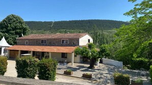 Un edificio de piedra con tejado de baldosas, rodeado de vegetación y vistas a una colina boscosa.