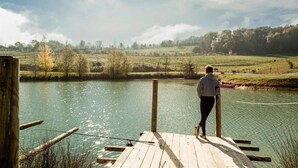 Un muelle de madera se adentra en una masa de agua tranquila, con una persona al final, contemplando un paisaje rural de campos y árboles.