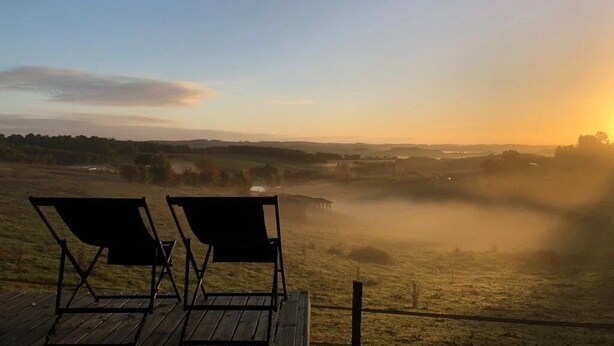 A wooden deck with two black lounge chairs overlooking a misty landscape during sunrise. - ZEcamping 4 étoiles familial piscine chauffée roulotte 9m2 2 pers (Estipouy)