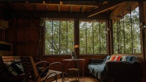Ein rustikales Blockhaus-Interieur mit Holzwänden, einem Schaukelstuhl, einem kleinen Tisch, einer Couch und großen Fenstern mit Blick auf Bäume.