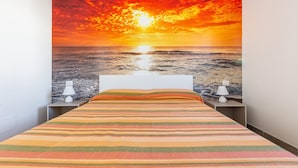Une chambre avec un lit, deux tables de chevet et une fresque murale représentant un coucher de soleil sur l’océan.