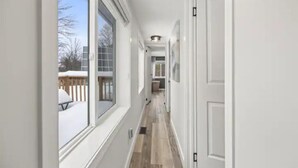A narrow hallway with white walls, a wooden floor, and large windows on one side.