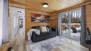 A cozy cabin interior with a wooden ceiling, a black sofa, a large window with a view of a snowy deck, and a painting on the wall.