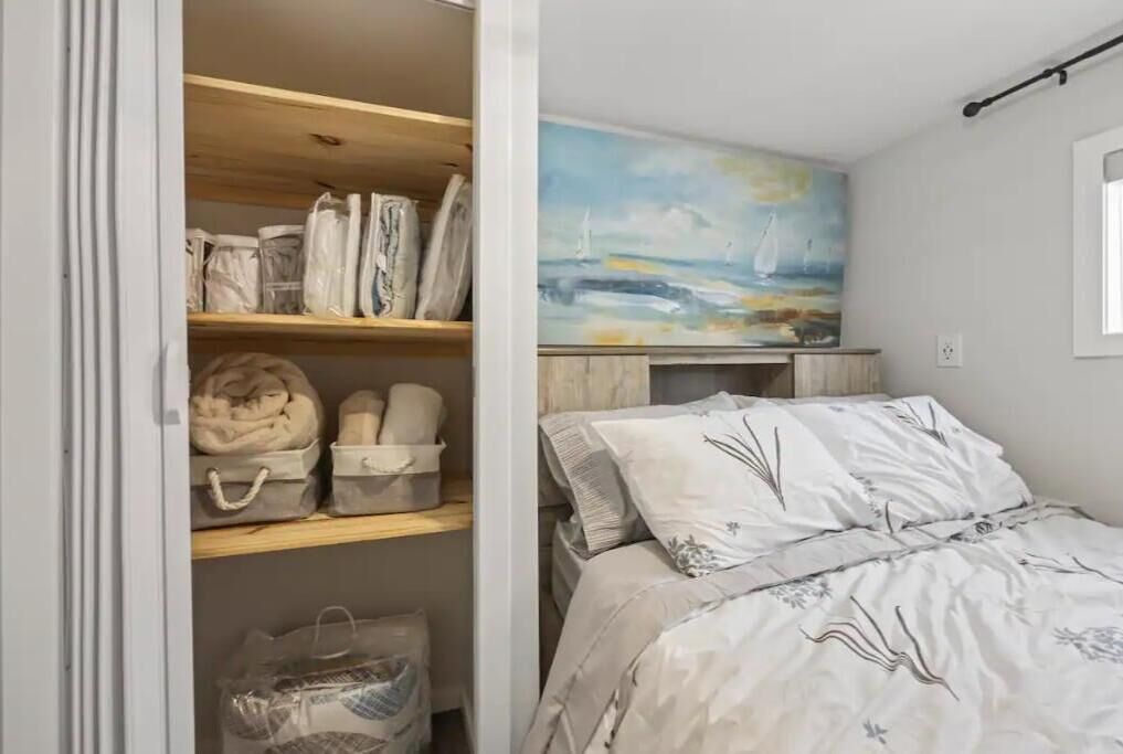 A bedroom with a bed, a painting of sailboats on the wall, and a closet with neatly folded clothes and bags.